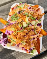 A top-down, close-up shot of loaded fries topped with slaw, sauce, and crispy onions, served in a polystyrene takeaway container on a wooden outdoor table.