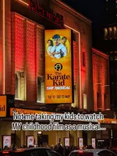 A night-time exterior shot of the Palace Theatre in Manchester, showcasing the illuminated promotional poster for 'The Karate Kid The Musical'.