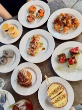 A flat-lay photograph of a variety of small plates, including salmon blinis, fried chicken, scones, and desserts, arranged on a dark table surface.