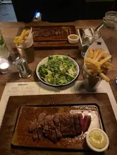A top-down shot of a steak dinner at a restaurant, featuring two steak boards, fries, and a salad. The lighting is dim and atmospheric, typical of a steakhouse setting.