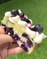 A close-up, handheld shot of a vegan blueberry and vanilla cake slice, showcasing the texture of the sponge, the cream topping, and the fresh blueberries. The image is taken outdoors against a soft, out-of-focus green background.