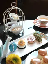 A high-angle, professional-style photograph of an elegant afternoon tea service, featuring a selection of delicate pastries, a macaron, and scones, set against a dark, sophisticated background.