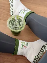 A lifestyle-focused shot featuring a matcha drink placed on a wooden floor between the legs of a person wearing pilates socks. The composition is clean and on-trend, effectively capturing a 'pilates and matcha' aesthetic.