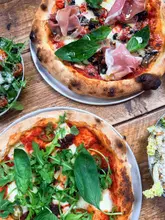A top-down, flat-lay photograph of two pizzas and two side salads arranged on a rustic wooden table, capturing a casual dining experience.