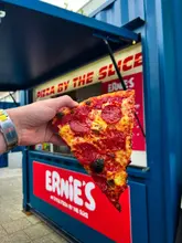 A close-up, eye-level shot of a hand holding a slice of pepperoni pizza in front of a blue shipping container venue. The focus is sharp on the pizza, with the venue's branding visible in the background.