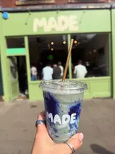 A bright, eye-level shot of a colourful smoothie or drink held in front of a cafe storefront named 'MADE'. The image uses a shallow depth of field to focus on the drink while keeping the venue branding visible in the background.