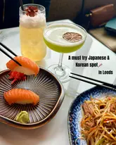 A high-angle, well-composed shot of a table spread featuring salmon nigiri being held with chopsticks, a noodle dish, and two cocktails, promoting a Japanese and Korean restaurant in Leeds.