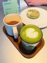 A bright, well-composed shot of a matcha latte and a cookie served on a wooden tray in a cafe setting. The lighting is natural and flattering, highlighting the vibrant green of the matcha and the texture of the cookie. The focus is sharp on the latte art, creating an inviting and professional aesthetic.