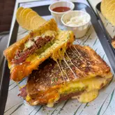 A high-quality, close-up food photography shot of a toasted sandwich, showcasing the melted cheese pull and crispy, golden-brown bread texture. The meal is presented on a branded tray with a side of crisps and two small pots of dipping sauces.