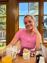 A bright, friendly portrait of a woman enjoying a breakfast of scones, jam, and cream in a sunlit cafe setting.