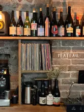 A stylish, atmospheric interior shot of a wine bar shelf, featuring a curated selection of wine bottles, a vinyl record collection, and coffee equipment against a rustic brick wall.