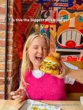 A promotional image featuring a creator holding a large, messy 'pickle burger' in front of a vibrant, graffiti-covered wall. The image is clearly intended for social media, focusing on the indulgence of the food item.
