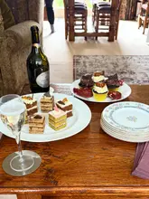 A casual, amateur-style photograph taken in a home setting featuring a selection of cakes, cupcakes, and a bottle of sparkling wine on a wooden table.