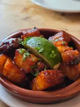A close-up, high-angle shot of a terracotta bowl filled with sticky, glazed pork belly bites, garnished with fresh parsley and a lime wedge, set against a wooden table background.