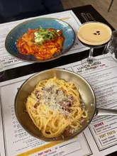 A top-down shot of a casual lunch spread at Zucchini Pasta Bar, featuring a bowl of carbonara and another pasta dish alongside an espresso martini, all served directly on top of the restaurant's printed lunch menu.