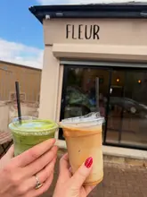 A casual, handheld shot of two iced drinks (a matcha latte and an iced latte) held up in front of a cafe called Fleur. The photo captures a simple, social moment, though the background is slightly cluttered with a fence and a car reflection.
