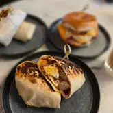 A well-composed, appetizing shot of a breakfast burrito cut in half, served on a dark plate, with a blurred background showing other menu items. The lighting is natural and flattering, highlighting the texture of the toasted tortilla and the filling.
