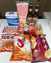 A flat-lay product haul featuring a variety of Japanese snacks, drinks, and condiments arranged on a plain white surface. The image showcases items such as Pocari Sweat, Kewpie mayonnaise, Asahi beer, and various Japanese snacks like Hello Panda and prawn crackers.