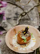A high-quality, close-up shot of a beautifully plated poached pear dessert, served on a rustic wooden plate in an outdoor, woodland setting. The dish is garnished with grains and edible flowers, reflecting a sophisticated, nature-inspired culinary experience.