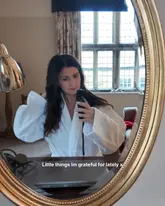 A mirror selfie of a woman in a white bathrobe, likely taken in a hotel room. The image captures a relaxed, lifestyle-focused moment, typical of travel or hotel stay content.