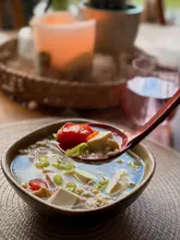 A warm, inviting close-up shot of a bowl of homemade egg drop soup, featuring silken tofu and tomatoes. The image uses a shallow depth of field to focus sharply on the ingredients held in a spoon, creating an appetizing and comforting visual.