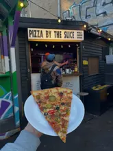 A first-person shot of a slice of pizza held up in front of a street food stall at a market. The pizza is topped with pickles, olives, and chillies.