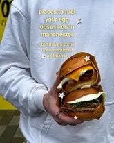 A close-up, eye-level shot of a person holding two stacked egg-based burgers against a plain yellow wall, featuring text overlay about egg-focused food spots in Manchester.