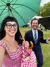 A candid, personal photo taken at a garden party event at Buckingham Palace. The image captures two individuals smiling under umbrellas, likely during an overcast day, highlighting the social and celebratory nature of the event.
