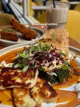 A bright, appetizing close-up shot of a brunch dish featuring poached eggs, greens, and flatbread, accompanied by a side of hash browns and an iced coffee. The natural lighting enhances the textures and colors of the food, creating an inviting atmosphere.