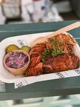 A close-up, high-quality shot of a street food meal served in a compostable container, featuring BBQ sausage, pulled pork, and red cabbage slaw, likely taken at an outdoor food festival.