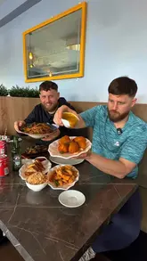 Two creators sit at a table in a restaurant, reviewing a spread of Chinese takeaway dishes including beef in black bean, salt and chilli chicken balls, and egg fried rice. They provide commentary on the taste and quality of the food throughout the video.