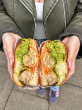 A high-quality, close-up shot of a meatball sandwich held in two hands, showcasing the cross-section of the ingredients including meatballs, pesto, and marinara on focaccia bread.