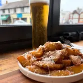 A close-up, eye-level shot of a plate of chips topped with grated cheese, positioned in front of a pint of beer. The setting appears to be a pub, with a window in the background providing natural light.