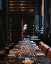 A sophisticated, moody shot of a long, elegantly laid dining table in a high-end restaurant, featuring a wine cellar in the background.