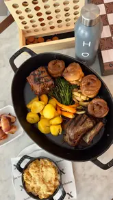 A couple enjoys a Sunday roast and plays various board games at The Hoxton, highlighting the venue's new 'Board Sundays' offering. The video features close-ups of the food, including a sharing roast, sides, and dessert, alongside shots of the couple interacting with games like Jenga and Connect Four in the hotel's lobby area.