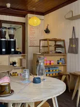 A candid, eye-level shot of a coffee shop interior, featuring a flat white on a wooden table in the foreground with the cafe's menu board and shelving visible in the background.