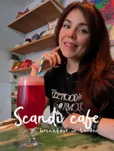 A casual, selfie-style photo of a creator sitting at a table in a Scandinavian cafe, holding a red drink. The image is clearly intended as a personal review or recommendation, featuring the cafe's interior and retail shelves in the background.