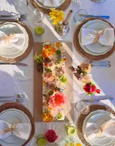 A high-quality, overhead lifestyle shot of a beautifully decorated rectangular cake adorned with fresh flowers, centered on a meticulously set dining table. The scene is bathed in warm, natural golden hour light, creating an inviting and elegant atmosphere.