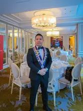 A formal portrait of a Maitre Hotelier standing in a sophisticated hotel dining room, likely during an awards ceremony or professional hospitality event.