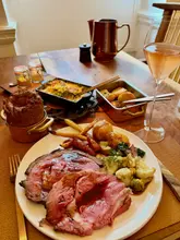 A high-quality, professional-grade photograph of a premium Sunday roast at The Connaught Grill. The image features a perfectly cooked beef rib, a large Yorkshire pudding, and seasonal vegetables, accompanied by a glass of rosé champagne. The lighting is soft and natural, highlighting the textures of the food, and the shallow depth of field creates a clean, focused composition.