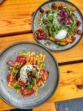 An overhead, flat-lay shot of two brunch dishes served on a wooden table. The top plate features avocado toast topped with a poached egg, microgreens, and what appears to be chorizo or sun-dried tomatoes. The bottom plate features a potato-based dish, likely hash browns, topped with bacon, a poached egg, spicy mayo, and pickled red onions.