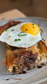 A short, static close-up video featuring a 'Highland Toastie' topped with a fried egg. The video focuses on the 'egg pop' moment where the yolk is broken, highlighting the texture of the dish.