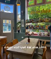 A bright, interior shot of a venue called Cultures in Saltaire, featuring a wooden table in the foreground and a large window display filled with wine bottles, looking out onto a street.
