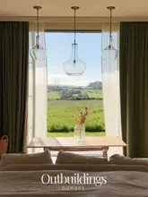A serene, symmetrical interior shot of a boutique hotel room in Dorset, featuring a large window overlooking a green field, framed by elegant curtains and hanging pendant lights.
