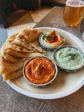 A top-down shot of a mezze-style platter featuring three bowls of dips, including hummus and a red pepper dip, served with warm pita bread, captured in a restaurant setting.