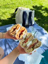 A first-person perspective shot of two people holding bagels on a picnic blanket in a park, with a tote bag in the background.