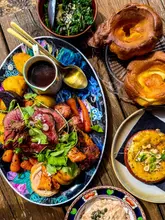 A top-down, sun-drenched shot of a Sunday roast spread featuring beef, pork, chicken, and various trimmings, served on a wooden table in an outdoor setting.