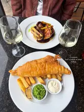 A high-quality, overhead shot of a dining table featuring a classic fish and chips dish and a chicken main, accompanied by two glasses of white wine. The image is well-lit and professionally composed, effectively showcasing the restaurant's food presentation and dining atmosphere.
