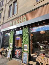 A straight-on, eye-level exterior shot of a cafe storefront named 'Mocha' on Maryhill Road, featuring outdoor seating, chalkboard menus, and a green foliage-decorated entrance.