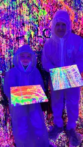 A vibrant, neon-lit montage showcasing an interactive splatter painting experience at Splash Art UK. The video features participants in protective suits using paintbrushes and water guns to create art on canvases and walls, highlighting the fun, messy, and creative nature of the activity.